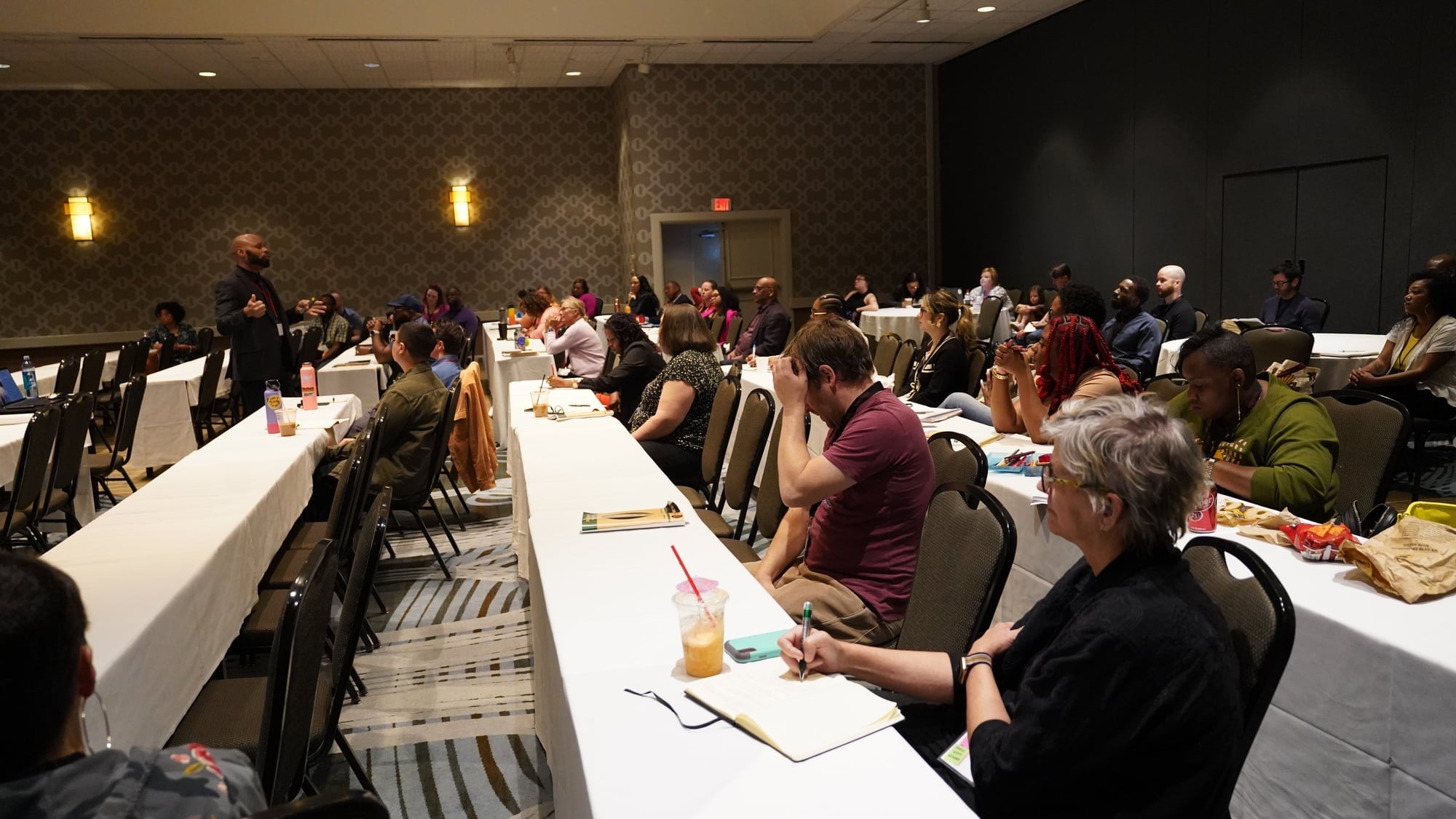 A hotel-ballroom training session with attendees seated at long tables taking notes — Dante King visible at left.
